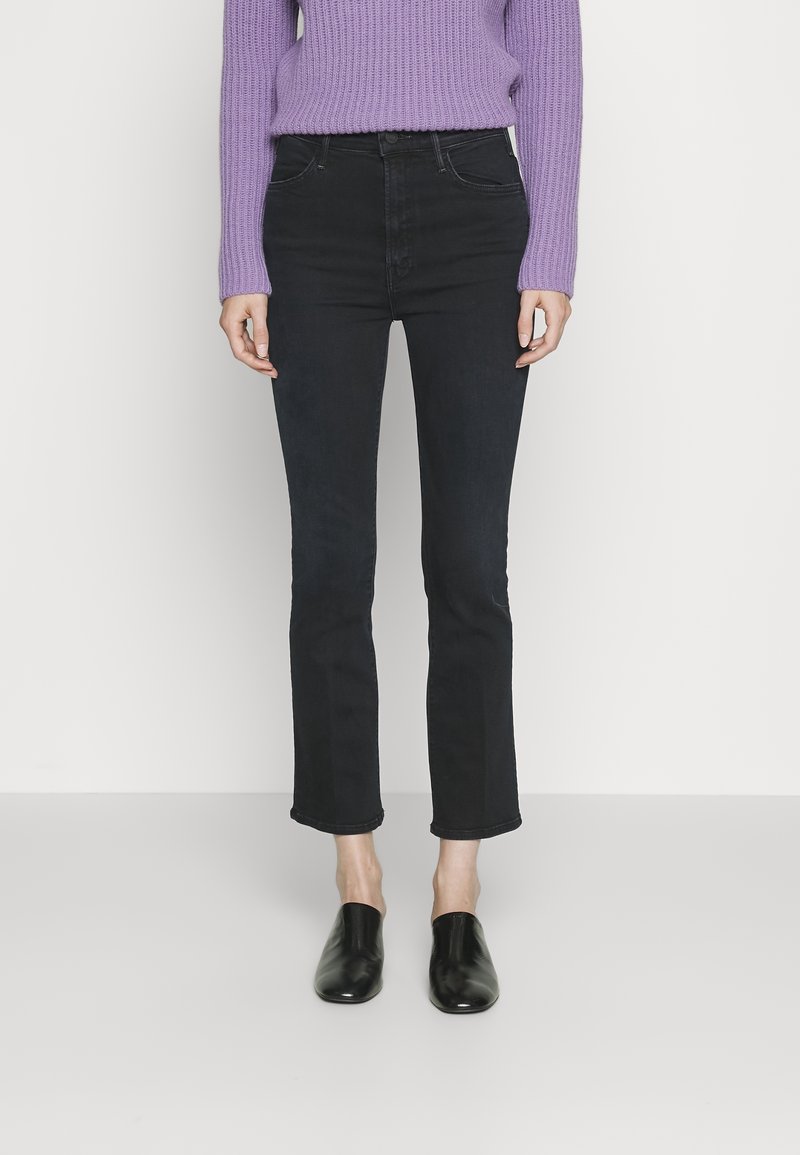Femme portant un jean noir taille haute à jambes droites, un pull en maille lavande et des chaussures slip-on noires, debout devant un fond clair uni.