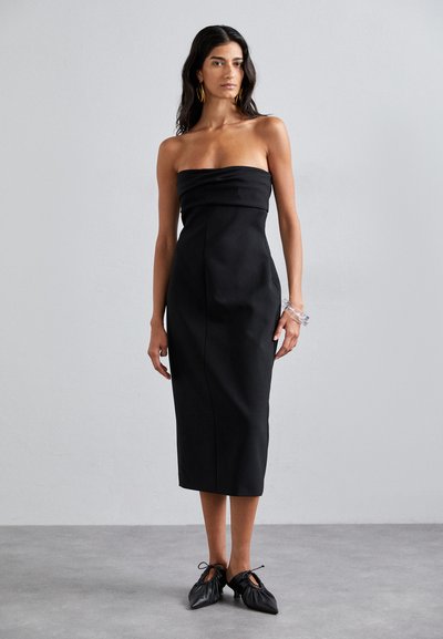 Schwarzes, trägerloses Midikleid mit gerafftem Oberteil und geradem Schnitt. Kombiniert mit schwarzen, gefältelten Schuhen und einem transparenten Armband am Handgelenk. Glatte Stoffstruktur.
