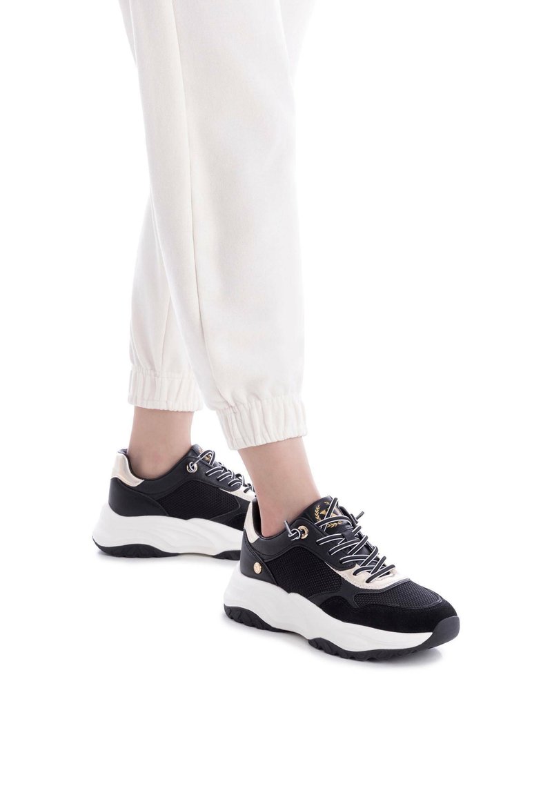 Zapatillas deportivas en blanco y negro con parte superior de malla, puntera redonda, suela blanca gruesa y cordones decorativos. Combinadas con unos pantalones jogger de color crema.