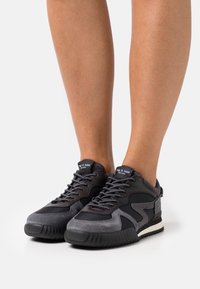Des baskets noires et grises présentent un mélange de textures lisses et en daim, avec un bout rond, une semelle rembourrée et des accents cousus.