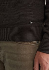 Brauner Strickpullover mit einem gerippten Saum und einem kleinen silbernen Logopatch auf der linken Seite, kombiniert mit strukturierten beigen Hosen.