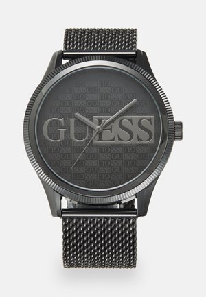 Montre noire avec un cadran gris foncé texturé arborant le nom de la marque "GUESS". Elle est dotée d'un bracelet en acier inoxydable maillé et de marqueurs d'heure argentés.