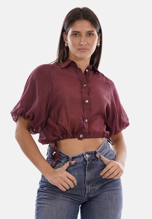 Donna con capelli castani lisci indossa una camicetta corta bordeaux trasparente con maniche a sbuffo e jeans blu a vita alta, in piedi con le mani vicino alle tasche.