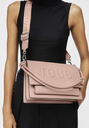 Borsa a tracolla in pelle rosa con logo "TOUS" impresso, manico intrecciato e tracolla regolabile. Caratterizzata da un design elegante e strutturato.