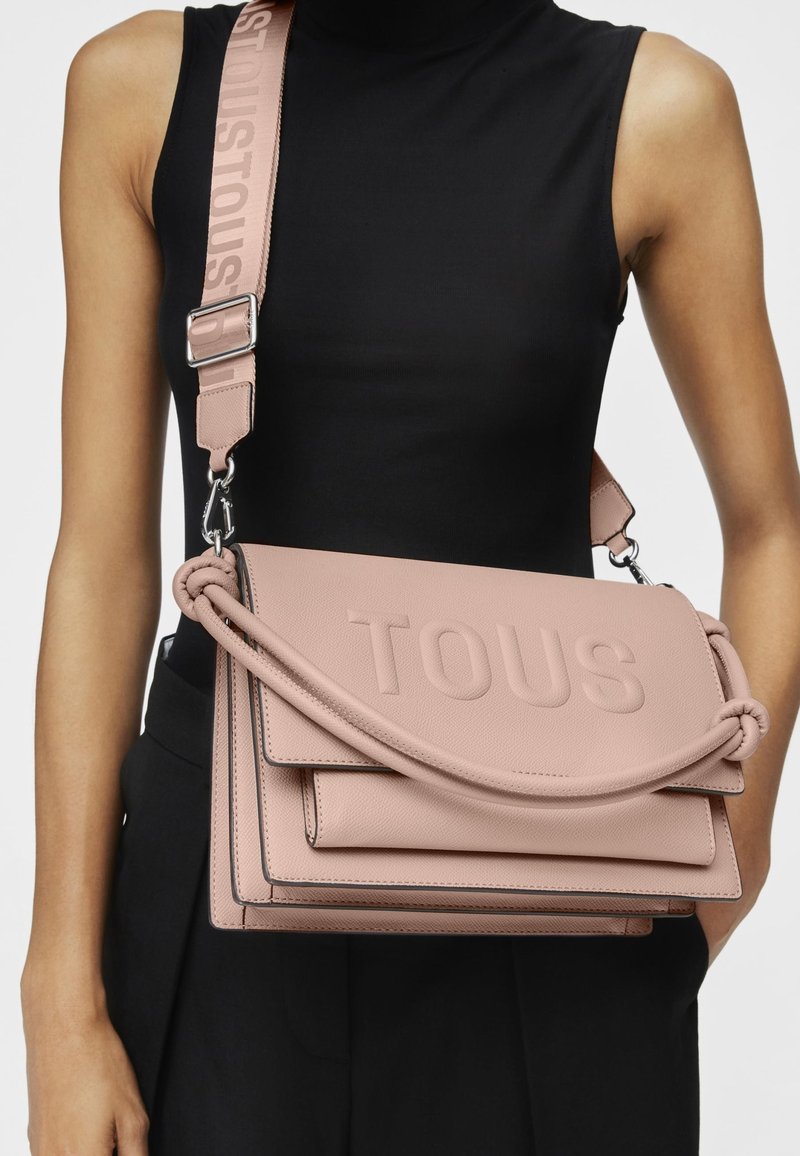 Różowa skórzana torebka crossbody z wytłoczonym logo "TOUS", plecionym uchwytem i regulowanym paskiem. Charakteryzuje się eleganckim, strukturalnym designem.