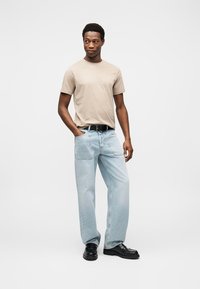 Μπεζ βαμβακερό μπλουζάκι, ανοιχτό μπλε τζιν σε φαρδιά εφαρμογή, μαύρη ζώνη και μαύρα παπούτσια. Το μοντέλο στέκεται με τα χέρια στις τσέπες σε ένα απλό φόντο.