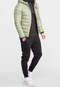 Person wearing light green padded jacket, black turtleneck, black joggers, white sneakers, and visible tattoos on hands and wrist.