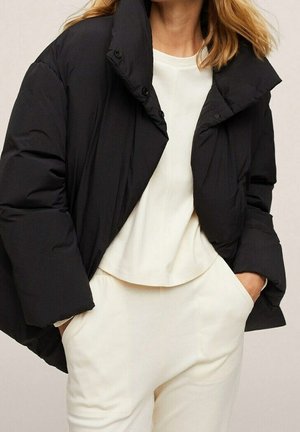 Kobieta nosząca czarną pikowaną kurtkę na białej bluzce z długim rękawem i białych spodniach, z rękami swobodnie w kieszeniach.