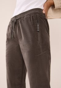 Joggers en tissu gris et doux avec une taille élastique, un cordon de serrage et une poche zippée sur le côté. Texture lisse avec une coupe décontractée et des jambes légèrement fuselées.