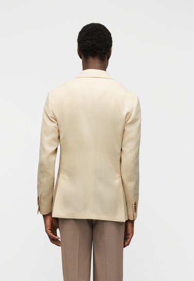 Άνδρας με ανοιχτό μπεζ σακάκι με υφή και παντελόνι ταουπ, στεκόμενος με την πλάτη προς την κάμερα μπροστά σε απλό γκρι φόντο.