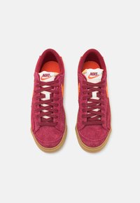 Paire de baskets Nike bordeaux avec swoosh orange, semelles en gomme, et languettes blanches arborant le logo Nike rouge, vues de dessus.