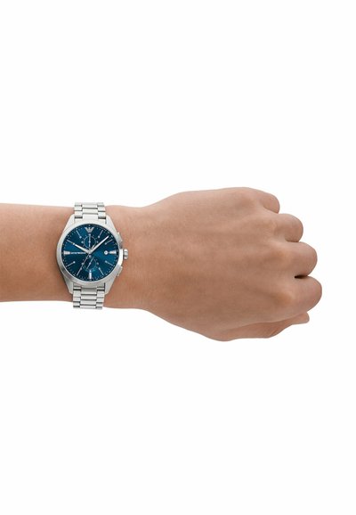 Montre-bracelet en métal argenté avec un cadran bleu, bracelet en acier inoxydable, boîtier rond et trois sous-cadrans, présentée sur une main.