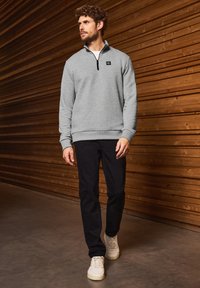 Felpa grigia con zip, in tessuto texturizzato, con polsini a coste e un piccolo logo, abbinata a pantaloni neri e sneakers bianche su uno sfondo di legno.