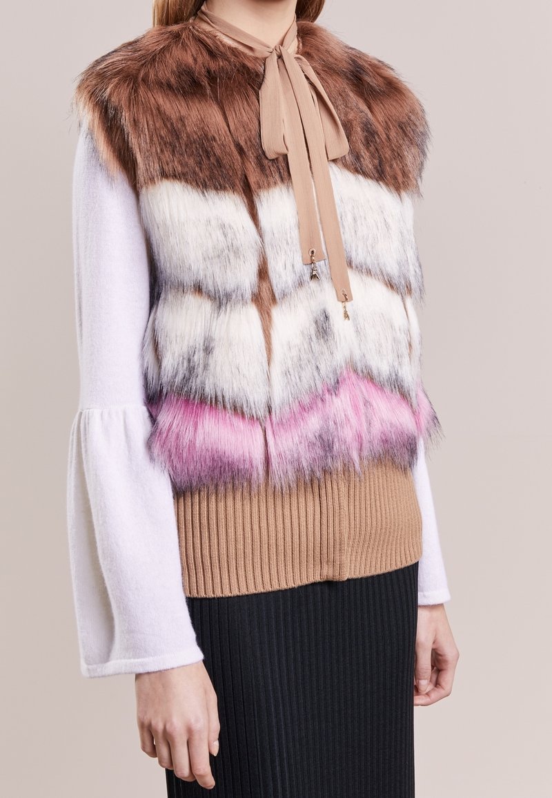 Faux pälsväst med en brun, vit och rosa chevronmönster, med en beige slipskrage, bärs över en ribbad brun topp och svart kjol.