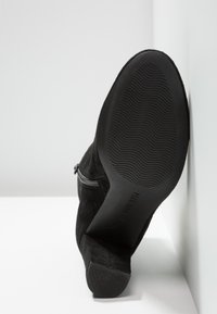 Botte cheville en daim noir avec fermeture éclair latérale, talon incliné et semelle en caoutchouc texturée vue de dessous sur fond blanc.