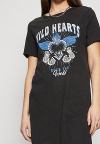 Musta t-paitamekko, jossa on graafinen kuvio sinisistä ja valkoisista sydämistä, siivistä ja kukkakoristeista, valmistettu pehmeästä puuvillamateriaalista.