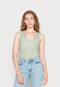Bodysuit verde sem mangas com um decote em V profundo, apresentando um intrincado bordado floral em prata e um design ajustado, combinado com jeans de cintura alta.