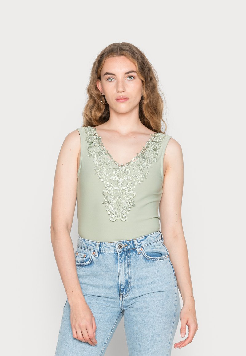 Bodysuit verde sem mangas com um decote em V profundo, apresentando um intrincado bordado floral em prata e um design ajustado, combinado com jeans de cintura alta.