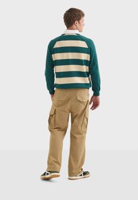 Zeleno-beige pruhovaná košile s dlouhým rukávem, khaki cargo kalhoty se bočními kapsami a černé tenisky s bílými detaily.