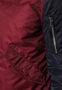 Burgundy- och marinblå vadderad jacka med en dragkedjficka, knappstängning på facket, slät textur och logotyp på axeln.