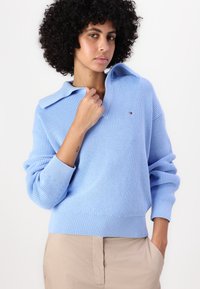 Pulover tricotat de culoare albastră deschisă, cu guler pliat, manșete cu aspect côtat și un detaliu mic cu logo. Materialul are un model texturat.