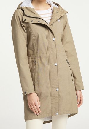 Imperméable beige à capuche avec des boutons-pression à l'avant, une poche zippée et des poignets ajustables, porté par-dessus une chemise rayée et un pantalon blanc.