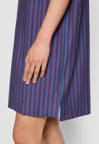 Vestido azul riscado com linhas verticais vermelhas, tecido suave, uma fenda lateral e um corte descontraído, mostrado com uma mão descansando na bainha.