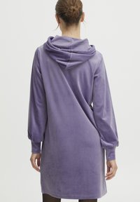 Lila, huva, långärmad klänning i mjukt, sammetsliknande tyg; har en lös passform, med puffärmar och en rundad rygg.