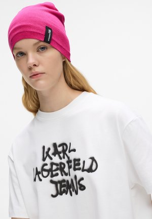 Rózsaszín kötött sapka bordázott szegéllyel, fehér pólóval párosítva, amelyen fekete spray-festett "KARL LAGERFELD JEANS" logó található.