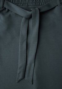 Tmavě zelená tkanina s hladkým povrchem, má široký pas a detail s uvázaným opaskem. Jednoduchý design s úhledným prošíváním.