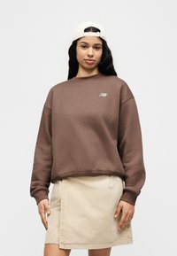 Καφέ φλις φούτερ με στρογγυλή λαιμόκοψη, διακοσμημένο με μικρό λευκό λογότυπο. Συνδυασμένο με ανοιχτό μπεζ τζην φούστα.