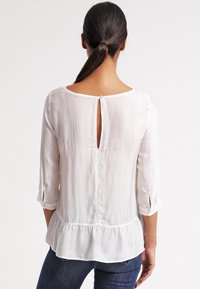 Blusa bianca leggera con maniche a tre quarti, dettaglio peplum e chiusura a oblò sulla schiena. Tessuto liscio e morbido.
