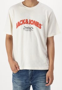 Cremfarbiges Kurzarm-T-Shirt mit dem roten "JACK&JONES"-Logo in fetter Schrift, das einen leicht lockeren Schnitt und eine weiche Baumwollstruktur aufweist. Getragen mit blauen Jeans.