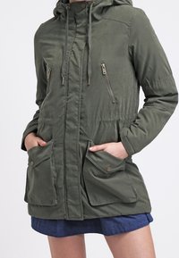 Olivově zelená bunda s kapucí na zip, se dvěma náprsními kapsami, elastickým pasem a nastavitelnými šňůrkami. Hladký textilní materiál.