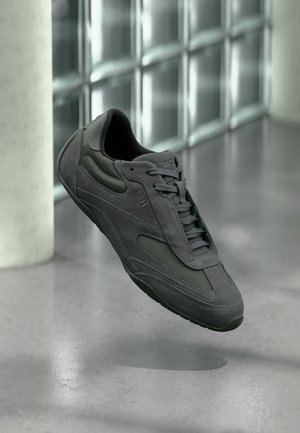 Baskets en daim et mesh gris foncé avec lacets et logo "BOSS" flottant au-dessus d'un sol poli près d'un mur en blocs de verre.