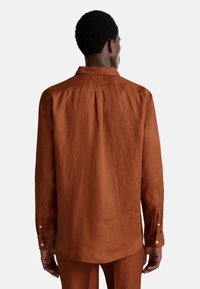Camicia in lino marrone con maniche lunghe, polsini con bottone e colletto classico. Presenta una vestibilità rilassata e dettagli con piega sul retro.