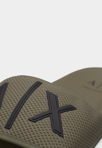 Κοντινό πλάνο του ιμάντα σανδάλιας σε λαδί πράσινο με ανάγλυφο μαύρο σχέδιο γραμμάτων "XV".