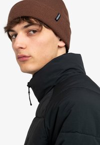 Zwarte pufferjack met een hoge kraag, gecombineerd met een geribde bruine beanie met een klein zwart logo-label. Glad materiaal en een aansluitend ontwerp.
