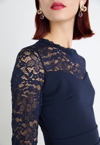 Marinblå klänning med spetslånga ärmar med blommönster, en figursydd bodice och en scalloped halsringning, som framhäver eleganta texturer och detaljer.