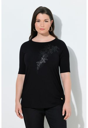 Frau mit langen dunklen Haaren, die ein schwarzes Oberteil mit Pailletten-Blumenmuster auf der Brust und eine schwarze Hose trägt, steht vor einer weißen Wand.