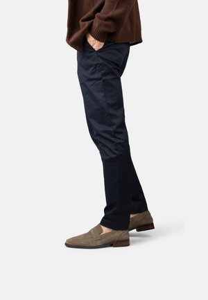 Mann trägt marineblaue, schmal geschnittene Hose, braunen Pullover und hellbraune Wildleder-Slipper, steht mit den Händen in den Taschen vor weißem Hintergrund.