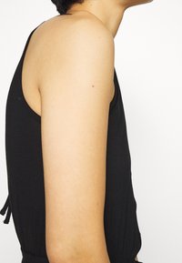 Czarna bezrękawnik z okrągłym dekoltem i otwartym tyłem, wykonany z miękkiego materiału o gładkiej fakturze.