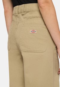 Beige wijde broek gemaakt van stevige canvas, met een achterzak met een merketiket. Het ontwerp heeft een hoge taille en een gestructureerde stof.