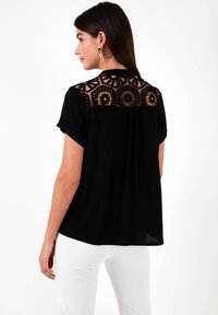 Maglietta nera con maniche corte e un design di pizzo sul retro con motivi circolari, completata da una vestibilità ampia e una leggera plissettatura.