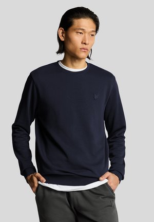 Junger Mann mit dunklen Haaren trägt einen marineblauen Pullover über einem weißen Hemd und dunkle Hose, steht mit den Händen in den Taschen vor einem einfarbigen Hintergrund.