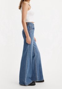 Jeans de mezclilla de tiro alto y de campana de color azul claro, con piernas anchas, que presentan costuras dobles y bolsillos traseros clásicos, combinados con una blusa blanca.