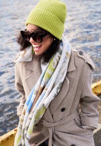 Cappotto beige doppiopetto con cintura, abbinato a una sciarpa leggera e fantasia. Berretto a coste verde e occhiali da sole neri oversize.
