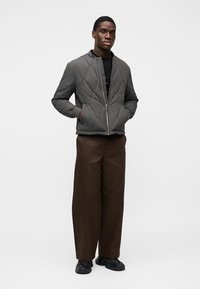 Γκρι μπουφάν bomber με σχέδιο καπιτονέ, φερμουάρ μπροστά, δύο πλαϊνές τσέπες και στρογγυλή λαιμόκοψη. Συνδυασμένο με φαρδιά καφέ παντελόνια και μαύρα αθλητικά παπούτσια.