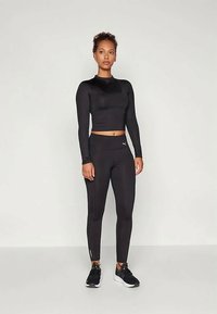 Fekete cropped hosszúujjú sportfelső, amelyet magas derékú fekete leggings-szel viselnek, textúrázott felülettel és logós részletekkel, fekete sportcipővel kiegészítve.