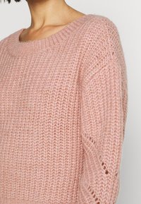 Jasnoróżowy sweter dziany z szerokim dekoltem. Posiada teksturowany wzór i drobne detale przy rękawach. Miękki, casualowy design.